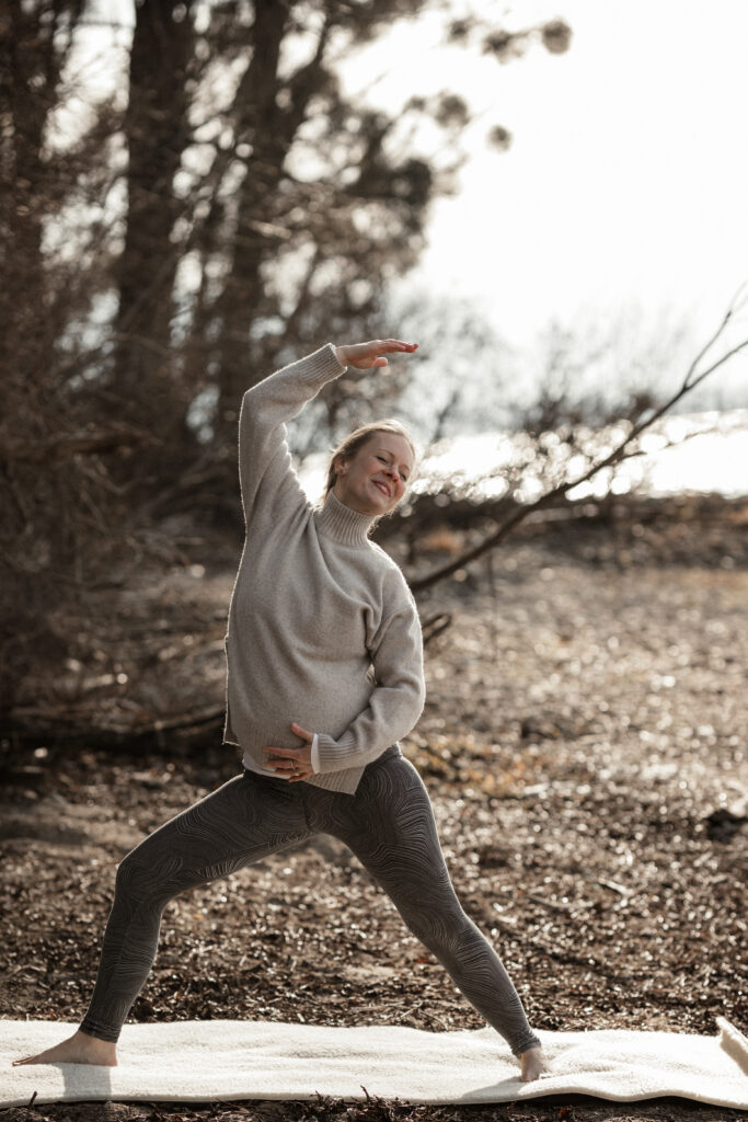 Eine lächelnde schwangere Frau in einem grauen Rollkragenpullover und gemusterten Leggings praktiziert Yoga auf einer weißen Matte im Wald. Ein Arm ist über den Kopf gestreckt, der andere ruht auf dem Babybauch.