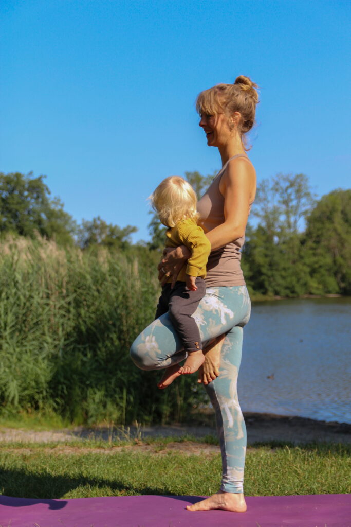 Rückbildungsyoga mit Baby – Mutter macht die Yoga-Baum-Haltung mit ihrem Kleinkind auf dem Arm am See.